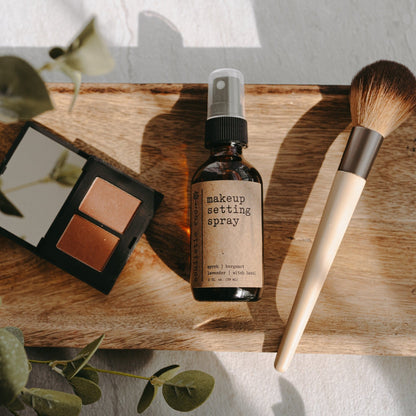 Makeup setting spray, brush, and eyeshadow palette on a wooden board with greenery.